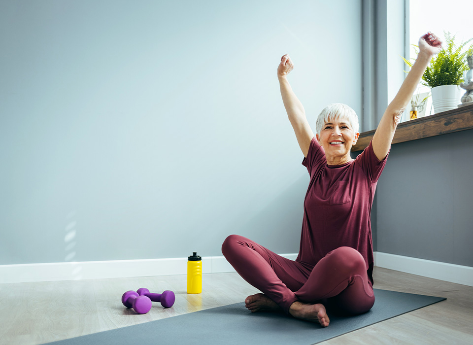 a woman sitting indoors on a yoga mat stretches her arms above her head