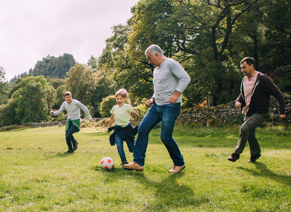 a boy plays soccer with three adult men of different ages outside on a lawn or park