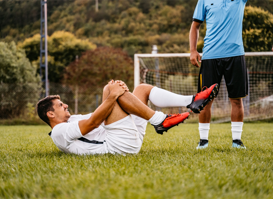a soccer player on his back on the ground, clutching his knee, while another player stands nearby