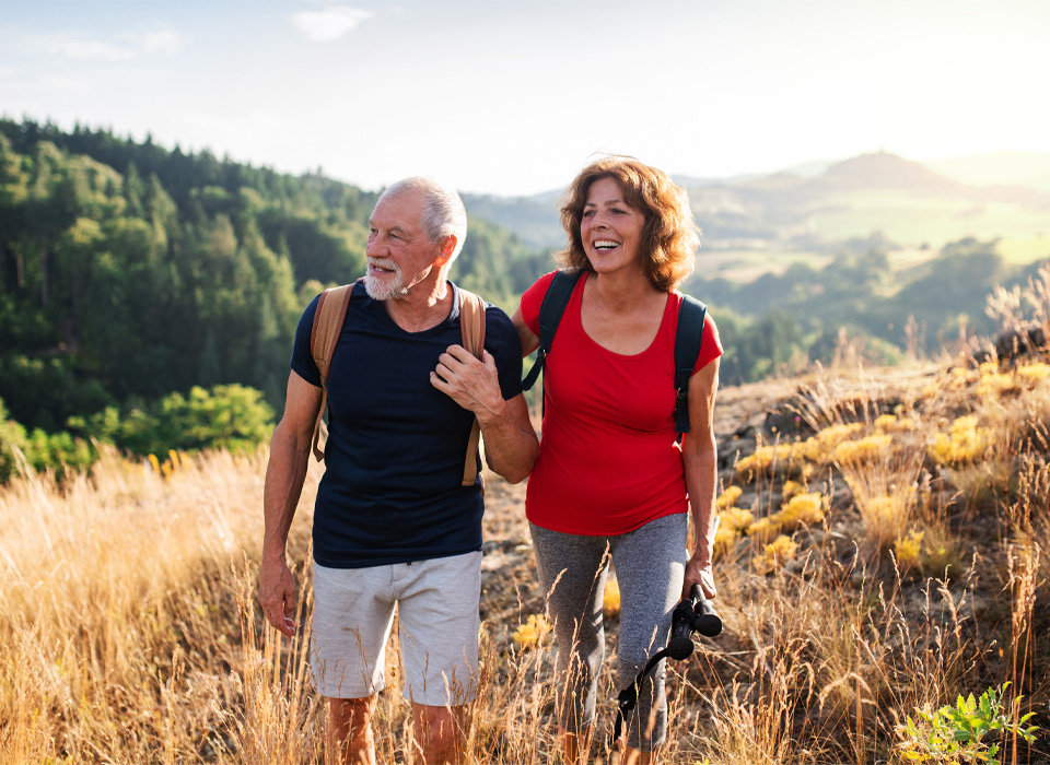 an older, active man and woman wearing backpacks hike on a sunny field on a mountainside