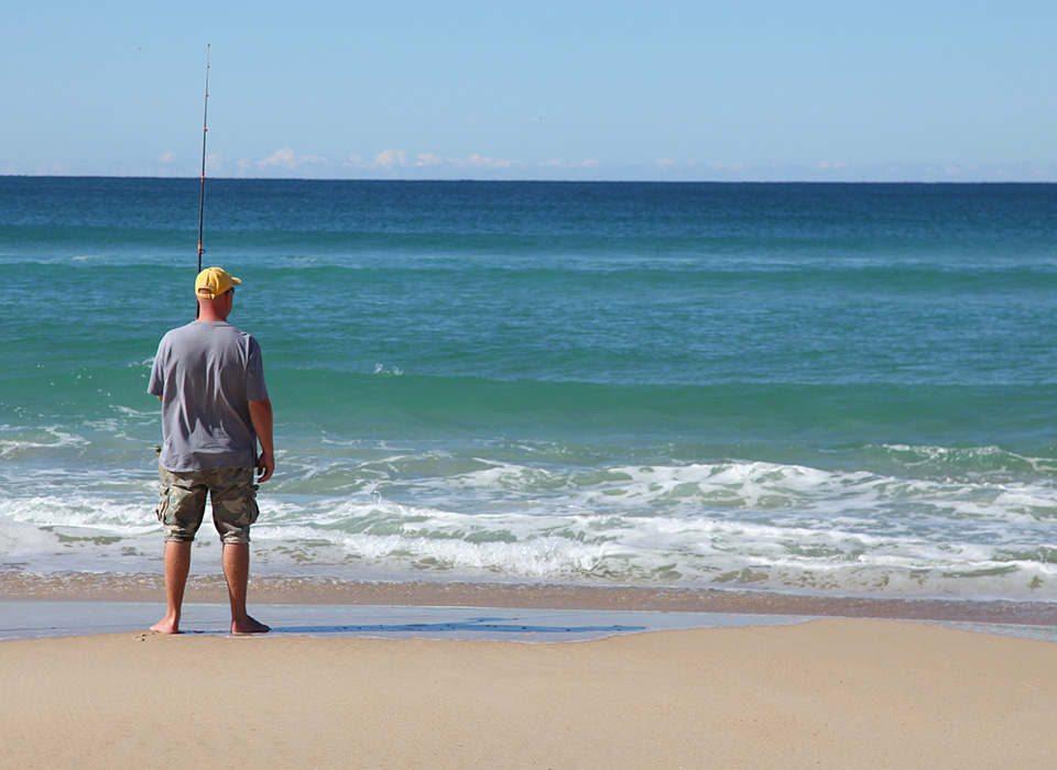 man standing on the sand at the edge of the ocean with his back to the camera, holding a fishing pole